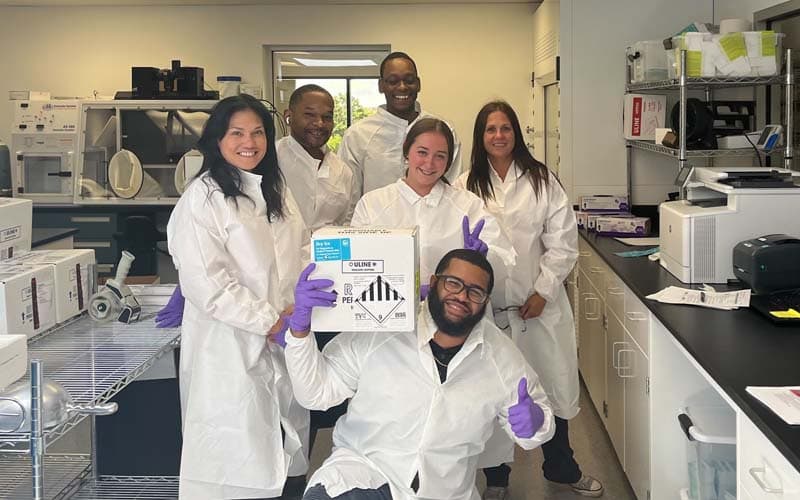 A group of scientists in lab coats and purple rubber gloves holding a box of stool samples and giving thumbs up